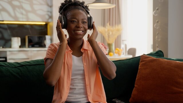 Relaxed happy smiling African American ethnic woman female girl lady homeowner businesswoman client headphones earphones listening music song sound dancing rhythm mp3 tune leisure indoors living room