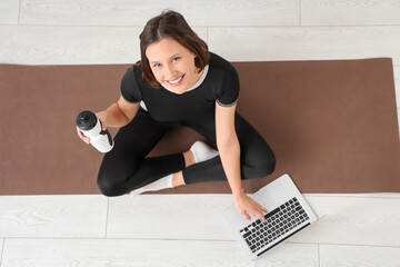 Sporty young woman with water bottle and laptop on fitness mat, top view