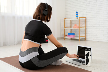 Sporty young woman in headphones using laptop on fitness mat at gym, back view
