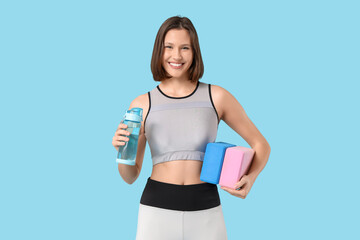 Sporty young woman with yoga blocks and water bottle on blue background