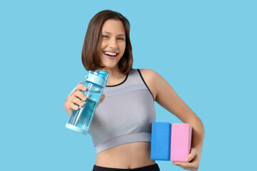 Sporty young woman with yoga blocks and water bottle on blue background