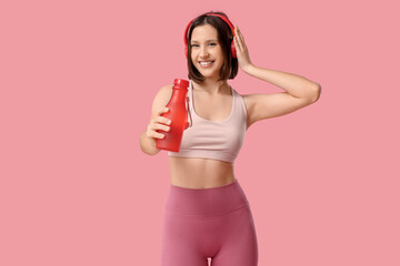 Sporty young woman in headphones with water bottle on pink background