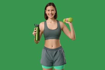 Sporty young woman with dumbbell and water bottle on green background