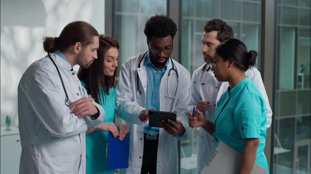 Group of five medical students diverse multicultural diagnostic team doctors men woman standing hospital lobby use tablet device nodding head agree prognosis healthcare rehabilitation discuss surgery
