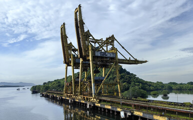 Abandoned yellow industrial port crane structure on a waterfront with lush green forest and cloudy...