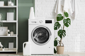 Modern washing machine with shelving unit, houseplant and detergents near white brick wall in...