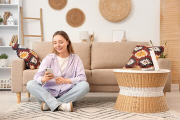 Beautiful young woman sitting on floor and using mobile phone in living room