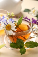 Aromatic herbal tea in glass cup, different flowers and mint on beige background, closeup