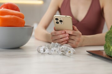 Woman using smartphone at white marble table with vegetables, kitchen scale and measuring tape...