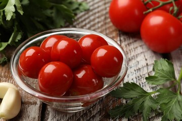 Tasty pickled tomatoes in glass bowl, parsley and garlic clove on wooden table, closeup