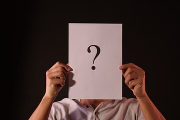 Man hiding behind paper sheet with question mark on dark background