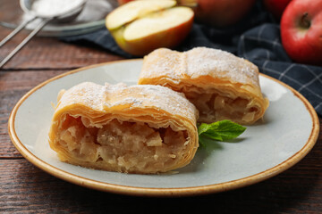 Pieces of tasty apple strudel with powdered sugar, mint and fruits on wooden table, closeup