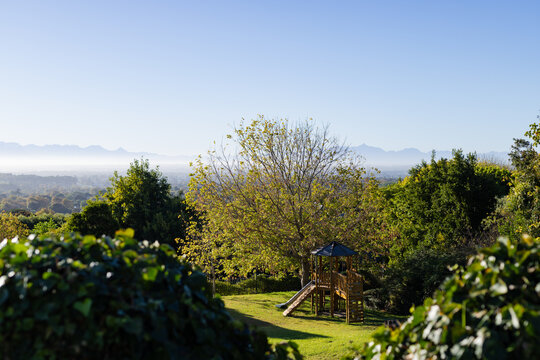Fototapeta Wooden playset with slide and ladder standing under tree on lawn, hedges framing distant mountains