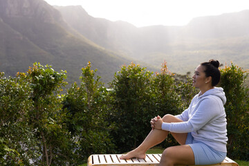 Asian woman sitting on wooden bench at mountain overlook among green shrubs and ridges, copy space