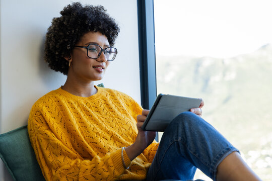 African American woman sitting by large window wearing yellow knit sweater using tablet, copy space