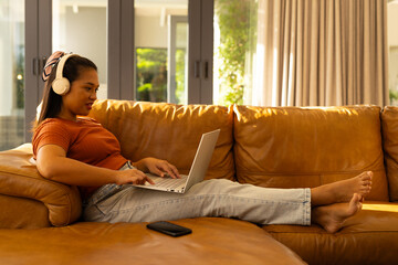 Asian woman typing on silver laptop at home wearing white headphones on tan leather sofa