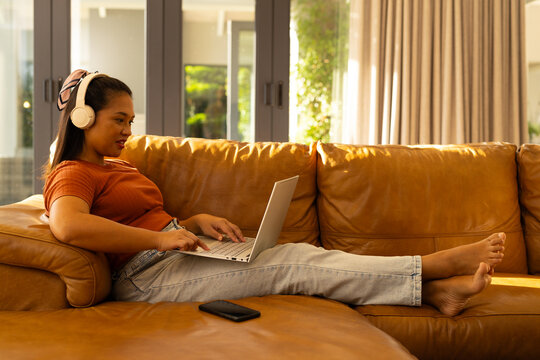 Asian woman typing on silver laptop at home wearing white headphones on tan leather sofa