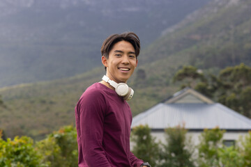 Asian man standing outdoors before mountain and metal-roofed house, wearing headphones around neck
