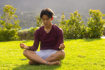 Asian man meditating cross-legged on green lawn in garden with trimmed shrubs and rolling hills