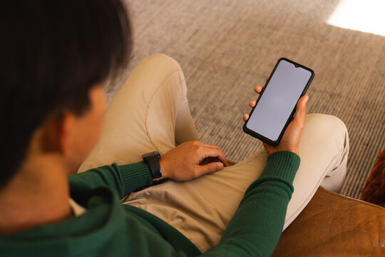 Asian male checking smartphone wearing sweater and smartwatch on sofa at home on carpet, copy space
