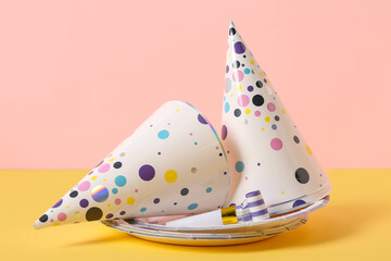 Composition with party hats, whistle and paper plates on yellow table near pink background, closeup
