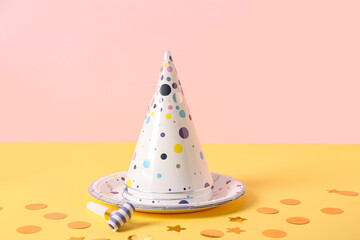 Composition with party hats, whistle and paper plate on yellow table near pink wall, closeup