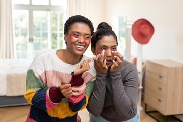 Diverse female friends applying red under-eye patches in bedroom near bed, hat stand with red hat