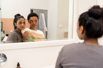 Diverse female friends applying under-eye skincare patches in home bathroom at vanity mirror