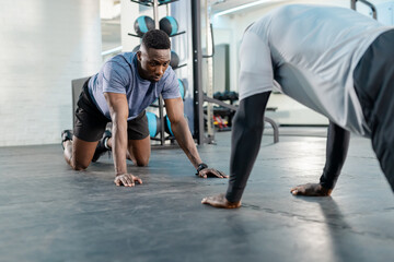 African American trainer and client performing bear crawl on gym mat beside medicine ball rack