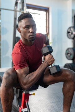 African American man lifting black hex dumbbell while performing one-arm row on red bench at gym