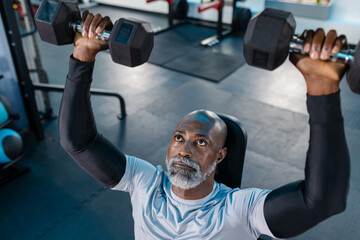 Senior African American man performing overhead dumbbell press on bench in gym with rubber floor