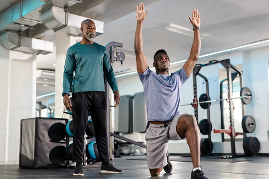 African American coach and client monitoring lunge form near squat rack on gym floor, copy space