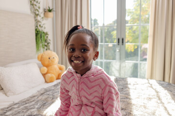 African American girl sitting on sunlit bed smiling brightly at camera with plush teddy bear