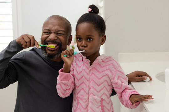 African American father brushing teeth at sink with daughter in pink pajamas near soap dispenser