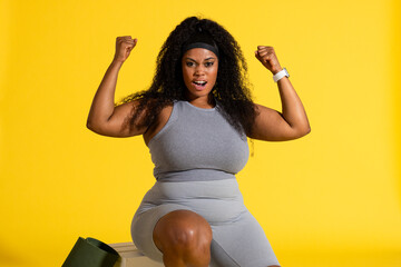 African American woman in gray sportswear flexing arms sitting on box in studio with yoga mat
