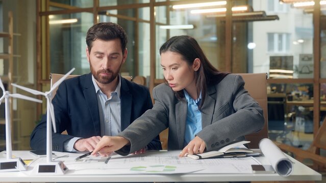 Japanese businesswoman inspector Caucasian businessman speak wind turbine model eco implementation renewable project document blueprint electricity engineering indoors office multinational meeting