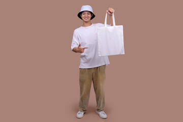 Ecology Concept. Asian Man Pointing with White Cotton Bag Over Beige Background.