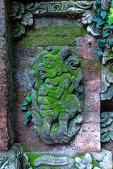 Moss-covered stone carving of a mythical creature adorns a brick wall in Ubud, Bali, Indonesia. The...
