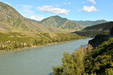 Obraz premium View of a wide river flowing through a valley surrounded by mountain ranges on a sunny summer evening.
