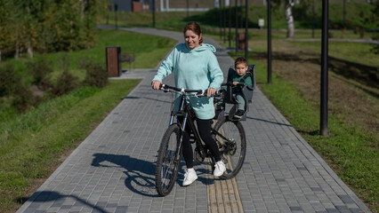 Caucasian woman riding bicycle with toddler son sitting behind her in child seat in park. 