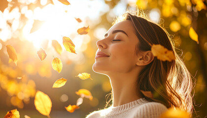 A young woman with closed eyes enjoys the autumn air as leaves fall around her in a sunlit park.
