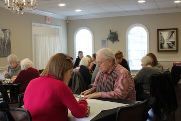 Group of people sitting at tables playing cards in a well lit room with windows and a chandelier