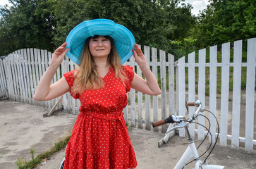Cheerful woman wearing a polka dotted red dress and a blue hat stands outdoors near a white fence...