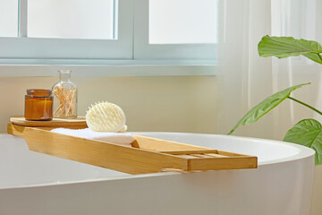 Bright bathroom view with wooden tray across bathtub, sponge, and jars. Soft light highlights the...