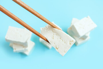 Piece of white tofu in wooden chopsticks on blue background close-up.