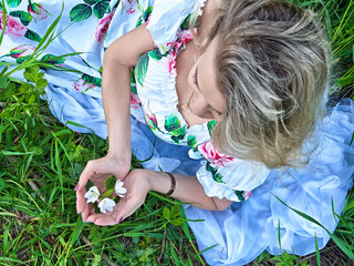 Blonde woman in floral dress poses with flowers while sitting in lush green grass during daylight