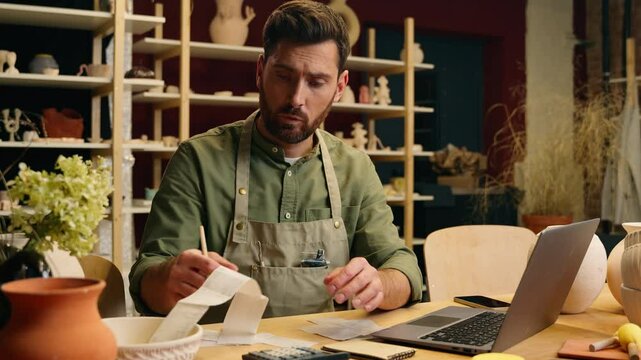 Frustrated despair Caucasian man guy businessman male small business owner potter crock working laptop calculating finance debt look receipt holding head gesture frustration bankruptcy inside workshop