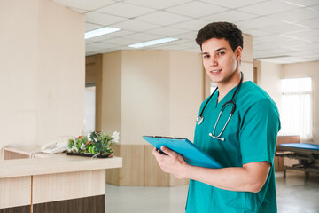 Young male nurse in green scrubs stands in hospital hallway, smiling while reviewing a patient’s...
