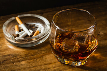 Glass of whiskey with ice beside an ashtray and cigarettes on wood