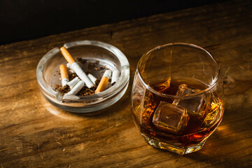 Glass of whiskey with ice next to ashtray and cigarettes on wood